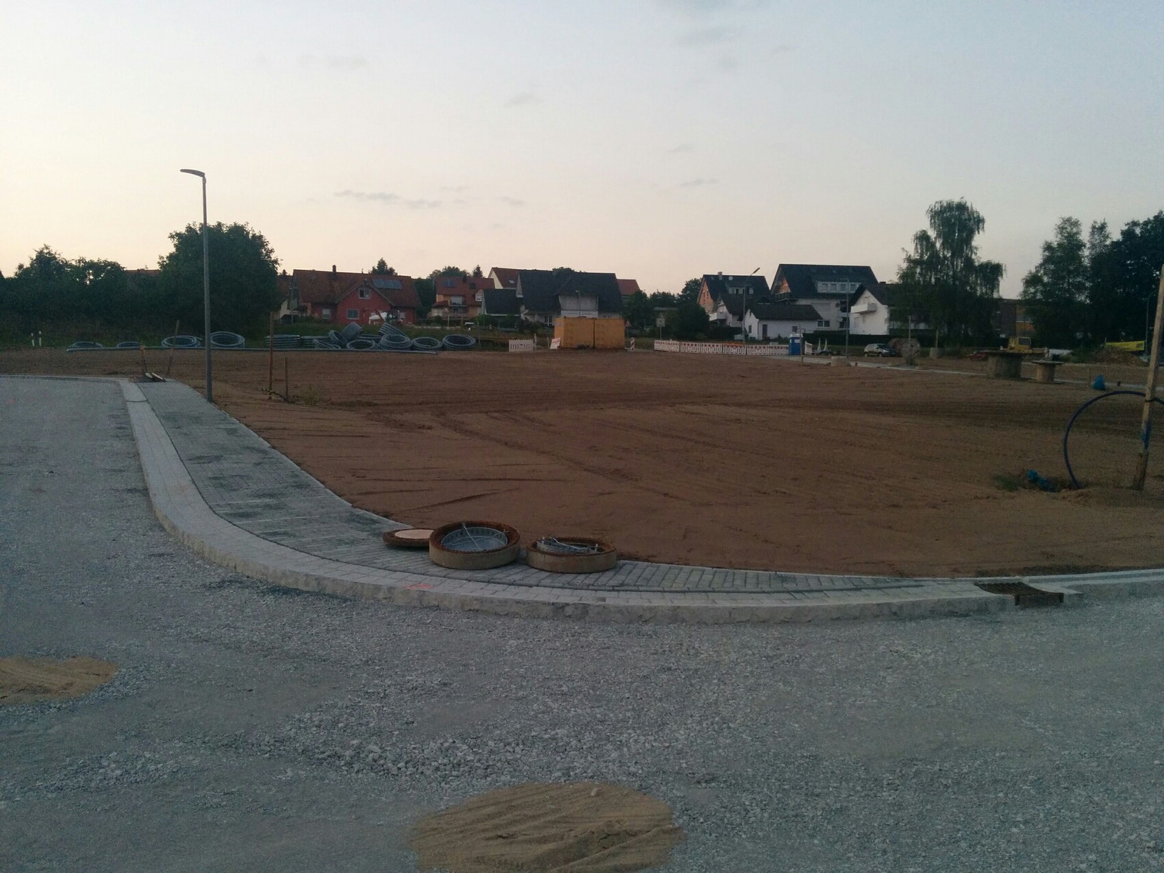 Baustelle mit Erdboden, sichtbare Rohre und runde Abdeckungen am Bordstein, Häuser im Hintergrund.
