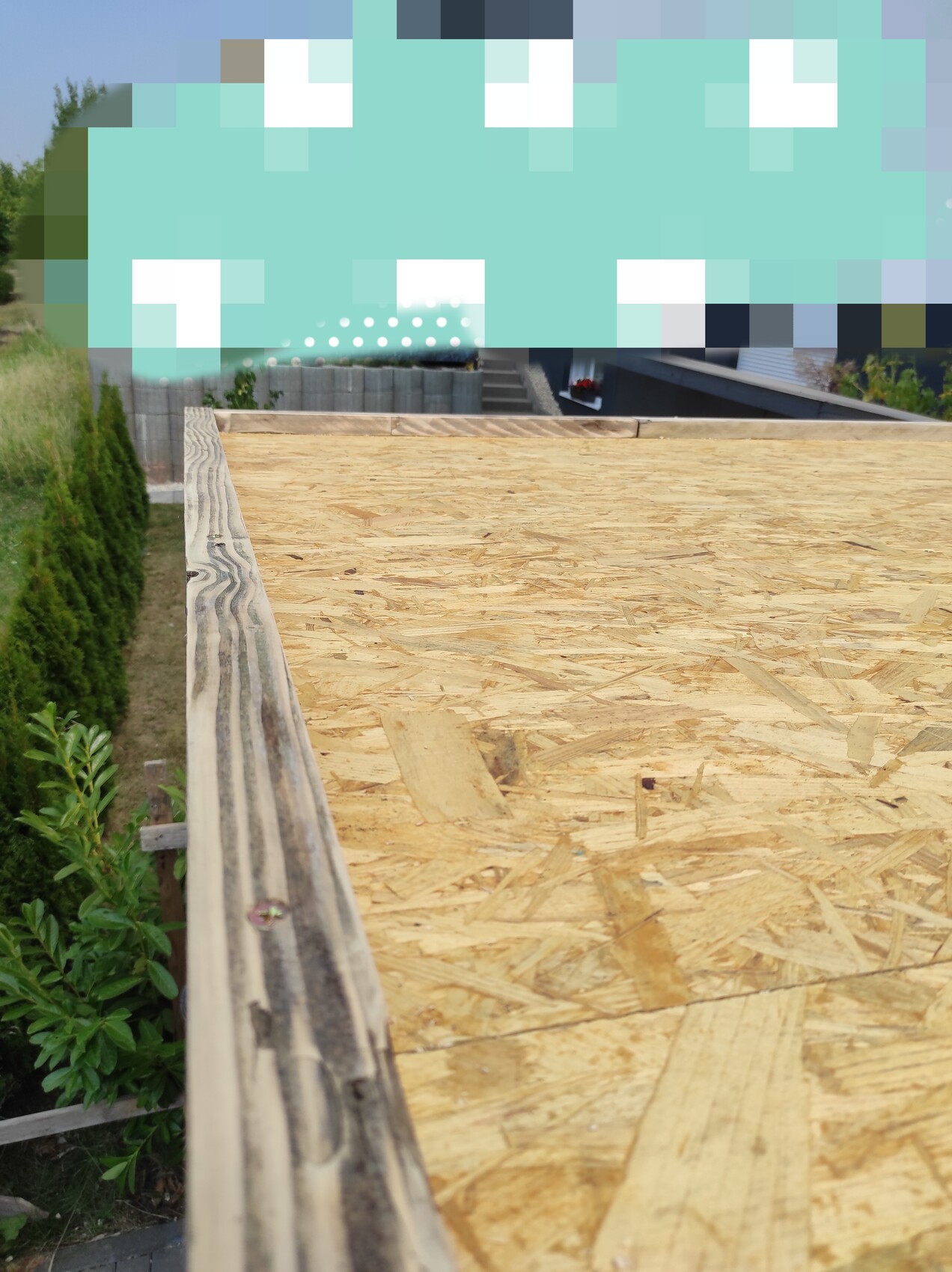 Blick auf offene Dachkonstruktion mit OSB-Platten, Holzkanten und Bauzaun, Gartenbereich sichtbar