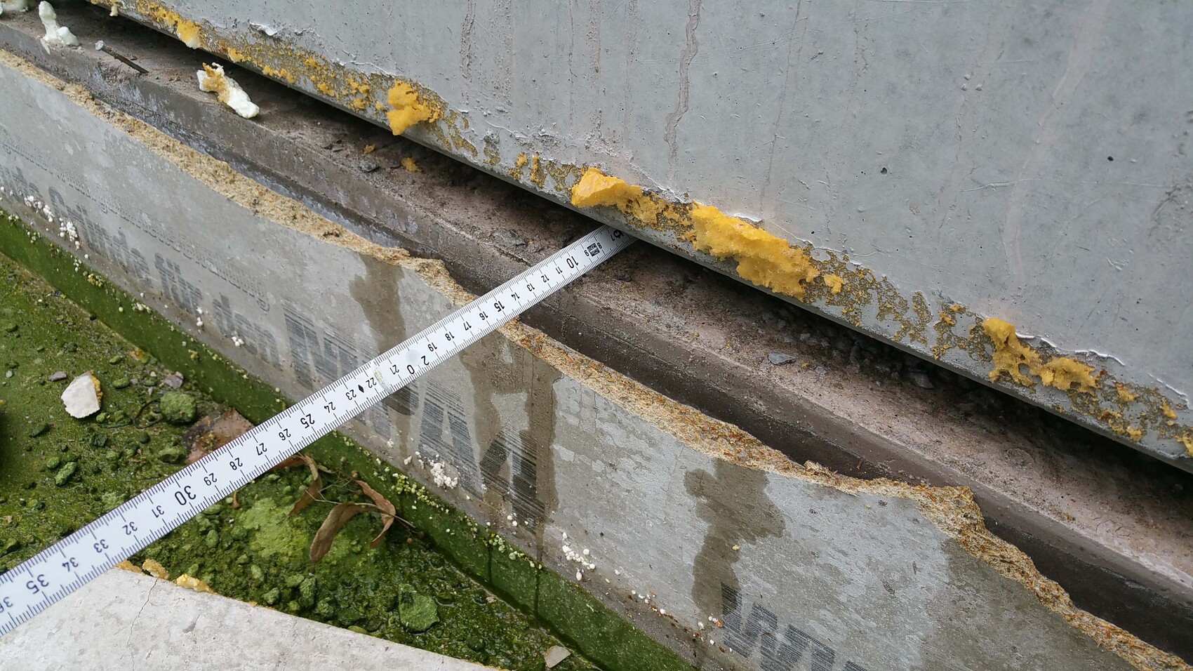 Betonfundament mit beschädigter Kante, gelbe Dichtmasse und Messband sichtbar, moosiger Untergrund
