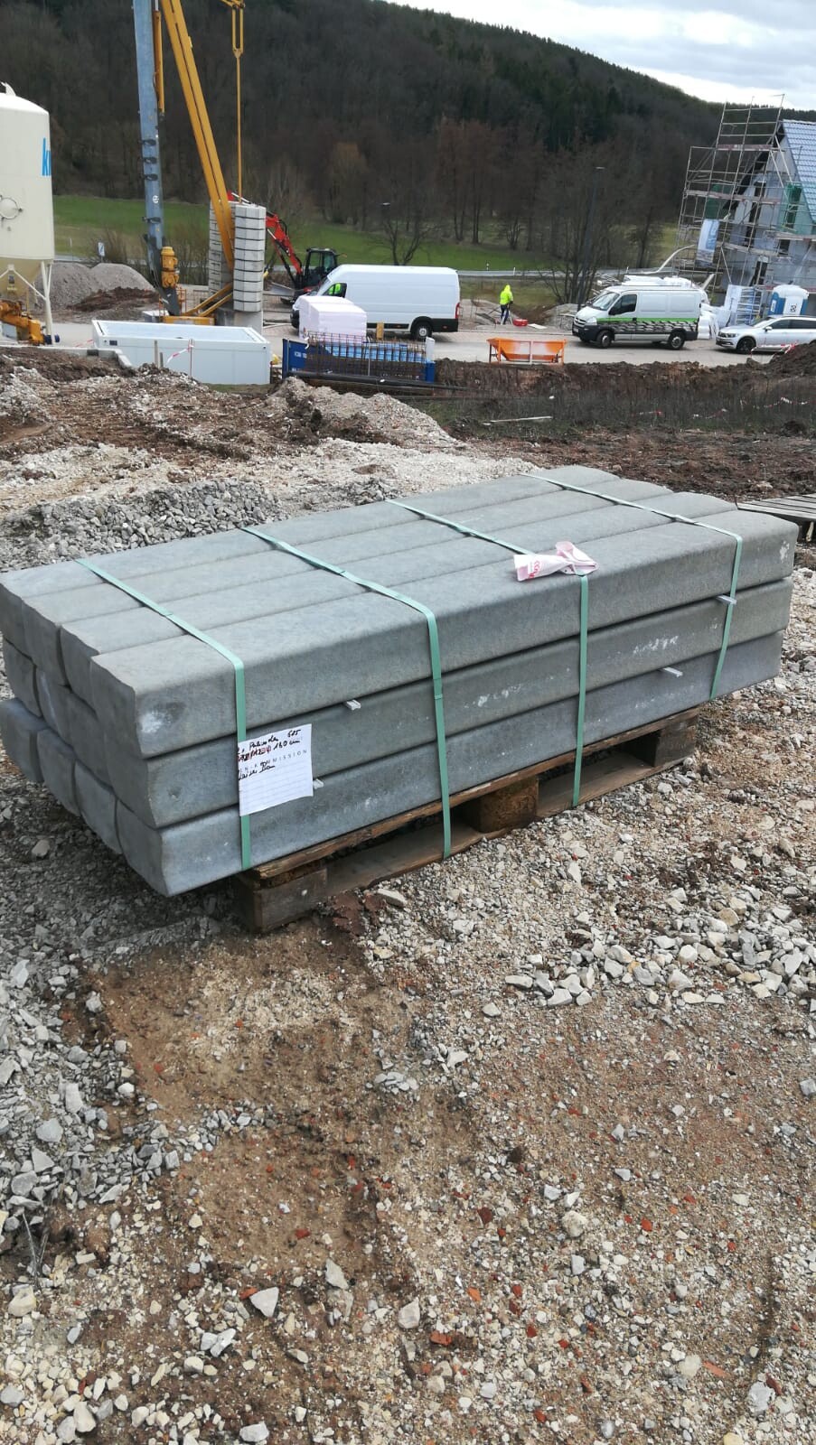 Gestapelte Betonblöcke auf Palette mit Spanngurten vor Baustelle, Fahrzeuge im Hintergrund