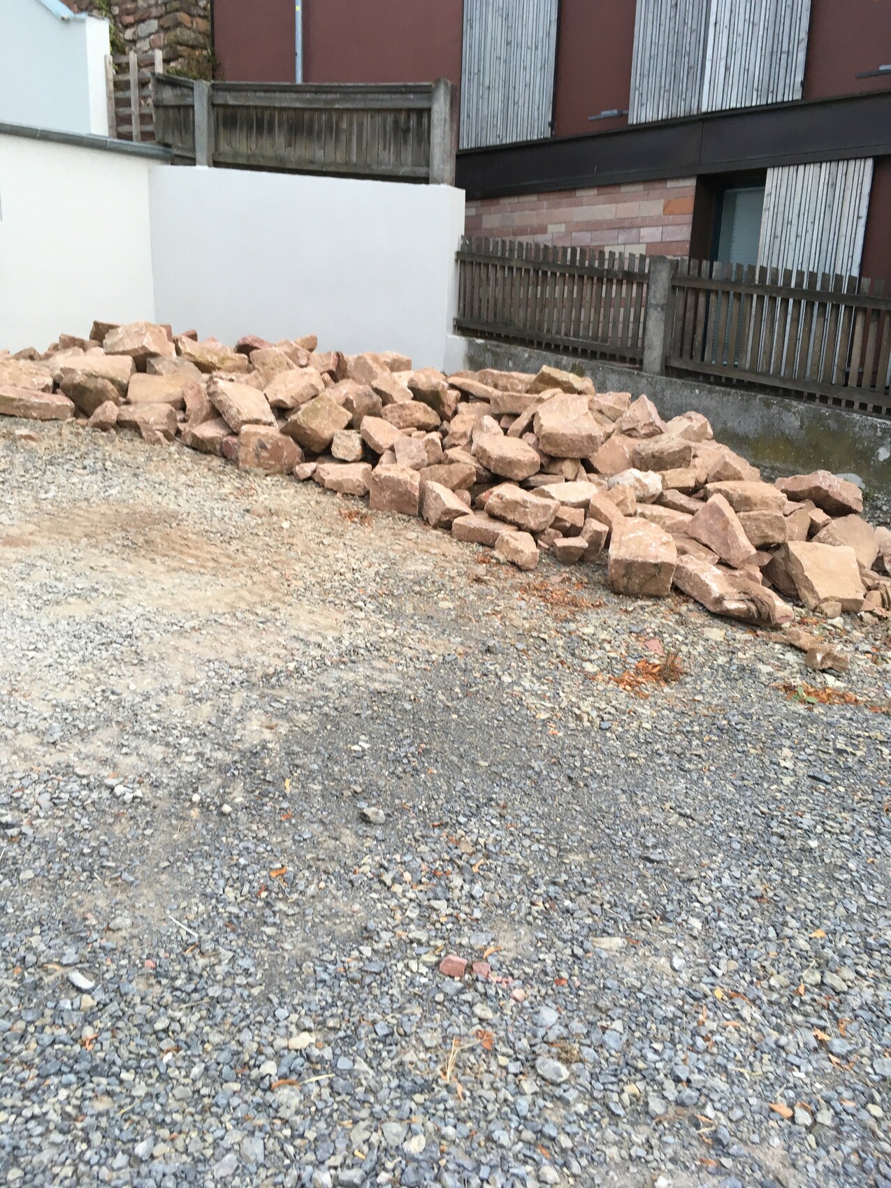 Baustelle mit Steinen, Kiesboden und Holzzaun vor Gebäude.