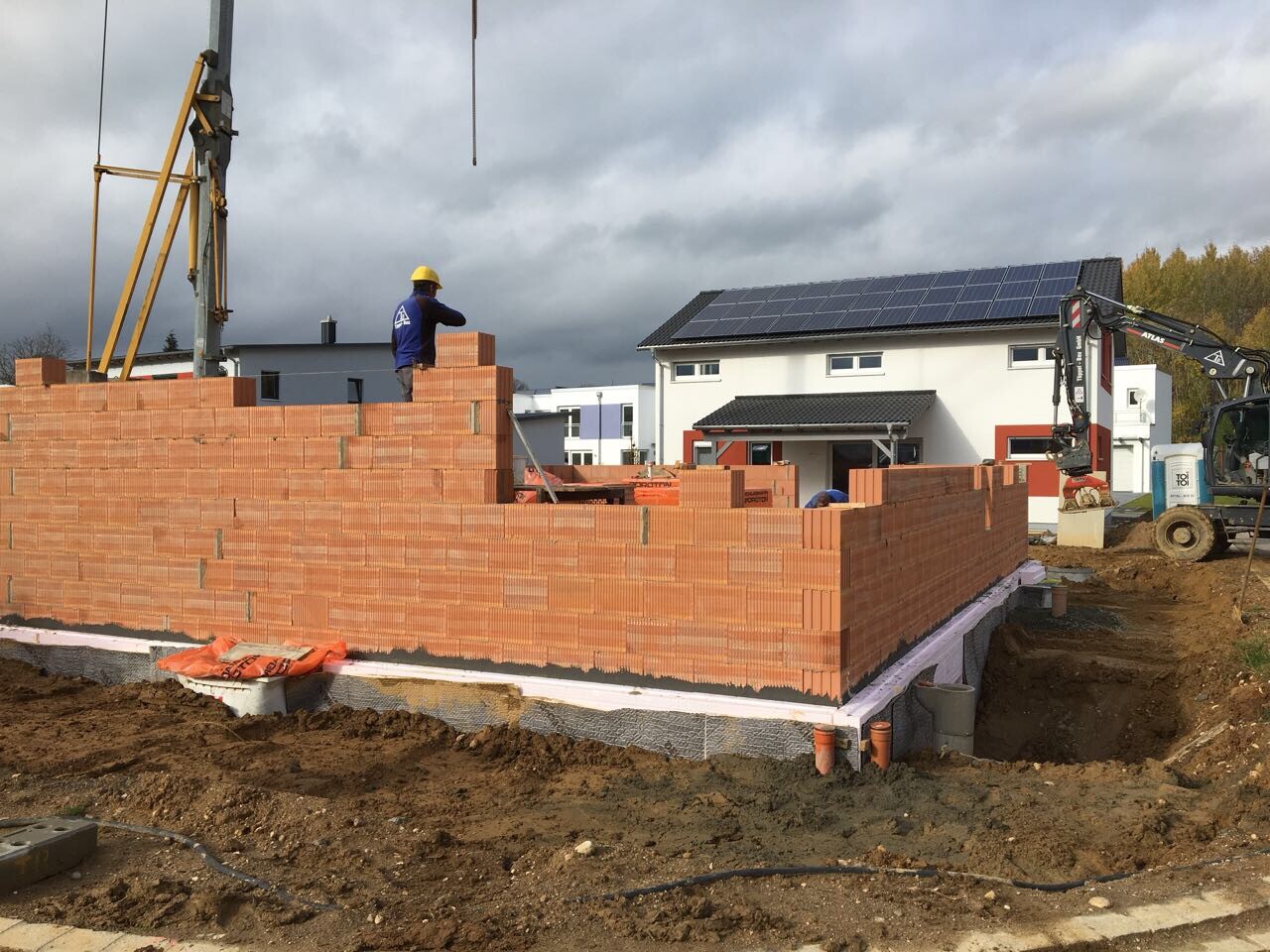 Baustelle: Maurer mit Gelbhelm mauert Ziegelmauer; Kran links, Haus mit Solarpanelen im Hintergrund.