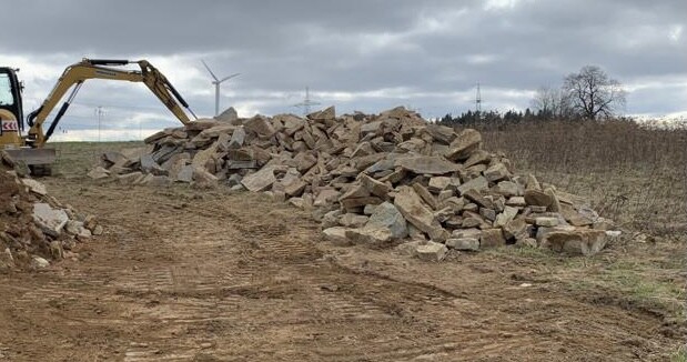 Bagger neben Steinhaufen auf Feldweg; Windräder im Hintergrund.