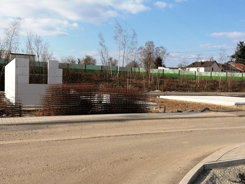 Baustelle an einer Straße: weiße Blockwand links, Bewehrung, Bäume und Häuser im Hintergrund.