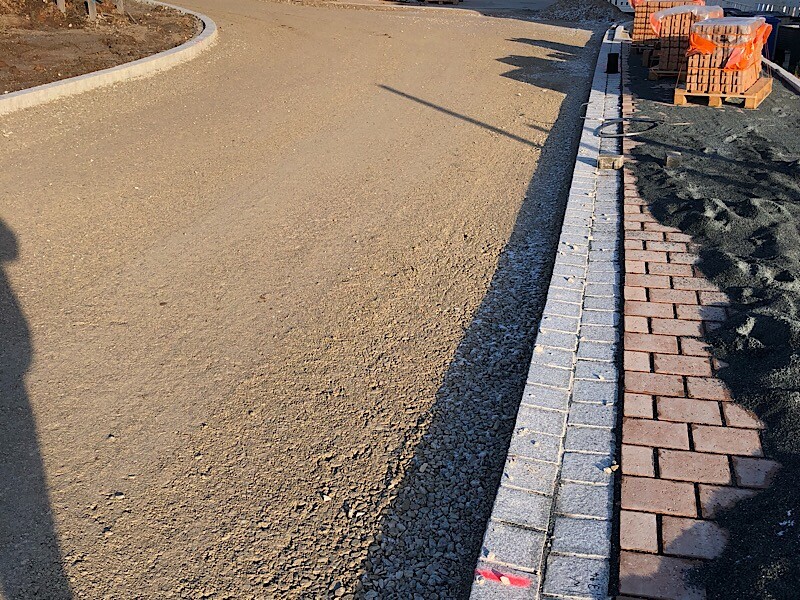 Außenbaustelle mit Kiesweg, Bordstein und Paletten mit Pflastersteinen.
