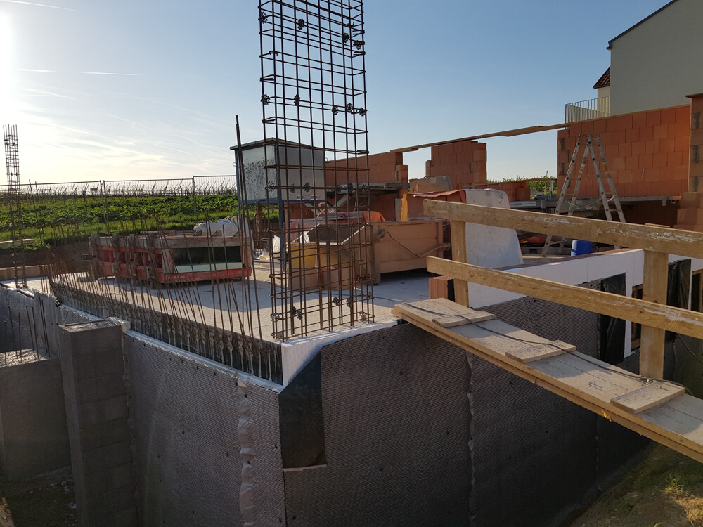 Baustelle mit Fundament, Stahlbewehrung, Backsteinwänden und provisorischer Holzbrücke.