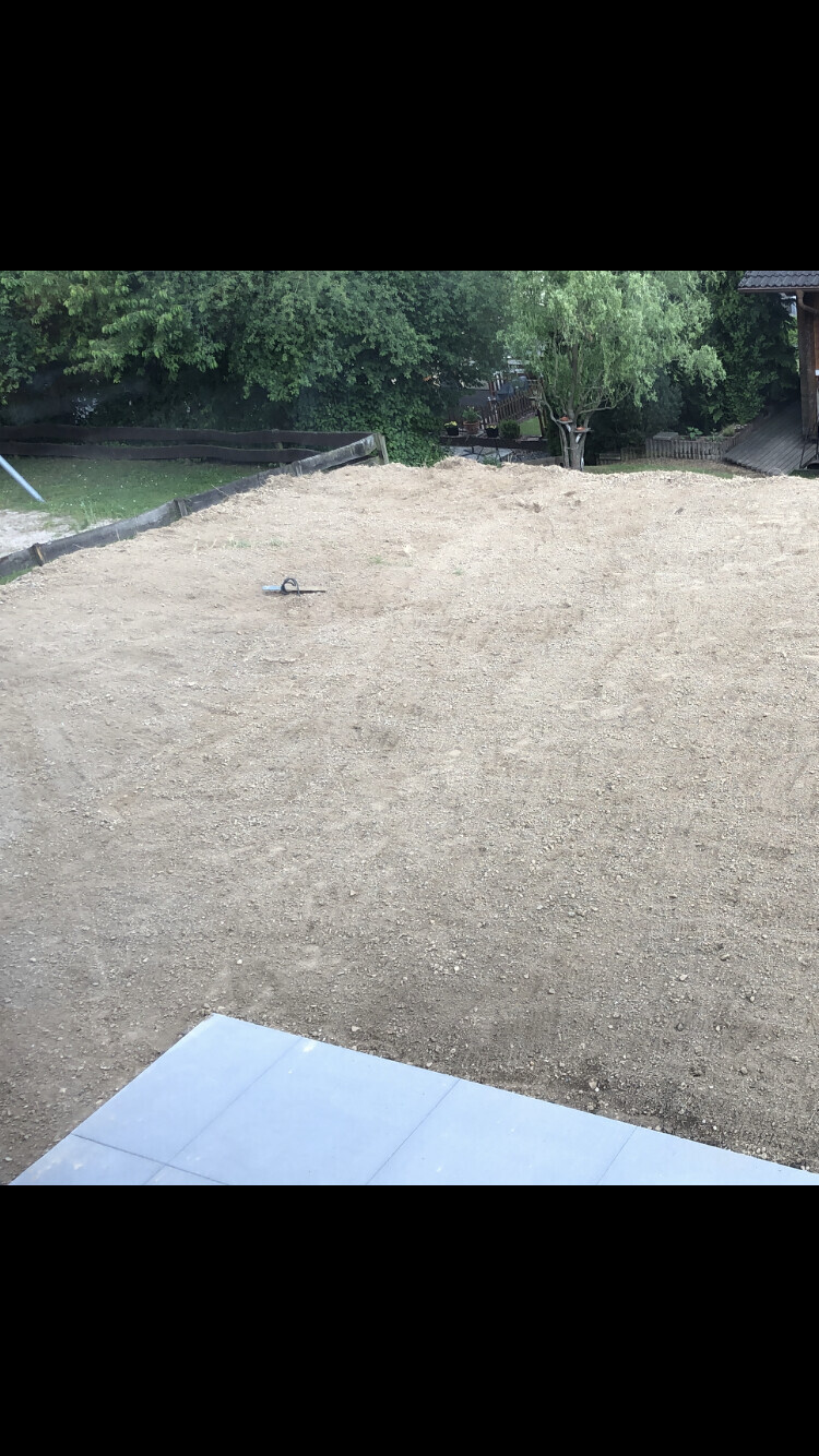Unbearbeitete Baustelle mit Kiesboden und freigelegtem Untergrund, Hintergrund Bäume und Garten