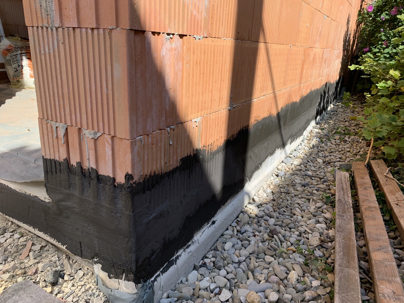 Außenwand aus roten Ziegeln mit schwarzer Dichtschicht am Sockel, Kiesgrund und rechts eine Holzbank.