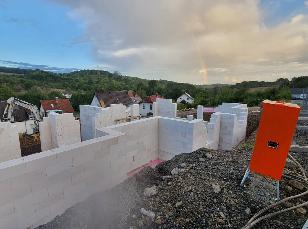 Baustelle mit weißen Betonblöcken, halbfertige Mauern, orange Verteilerkasten; Regenbogen am Himmel