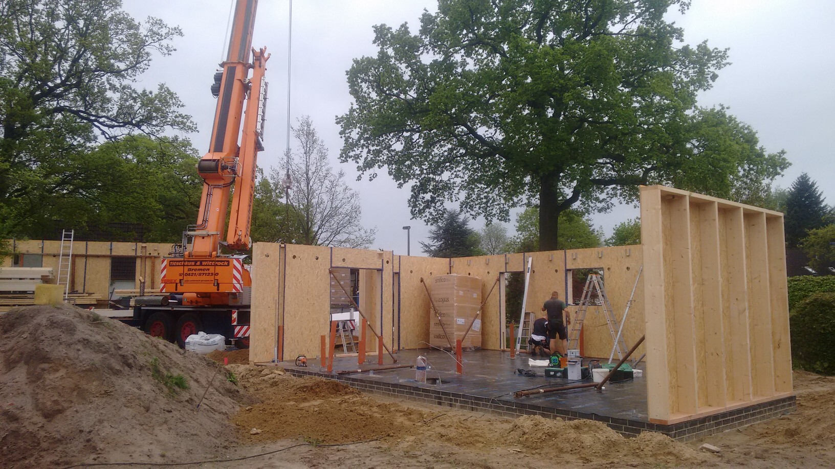 Baustelle: Kran hebt Holzrahmen-Wand, Bauarbeiter arbeiten am Fundament.