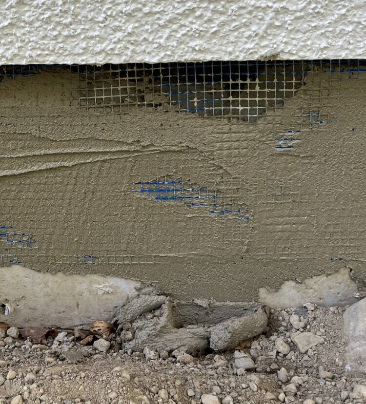 Verputzte Wand mit blauem Drahtgitter unter Putz; Geröll am Fundament.
