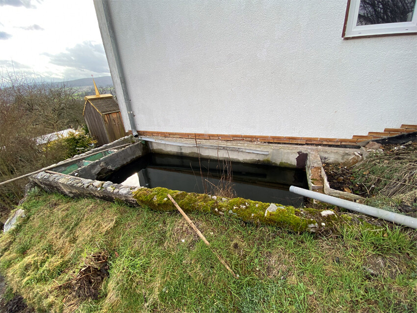 Offenes rechteckiges Wasserbecken neben dem Haus, von Moos überwachsen, mit Rohr daneben.