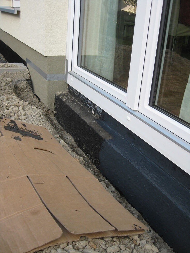 Außenansicht einer Baustelle am Fundament mit Fenstern und schwarzer Abdichtung.