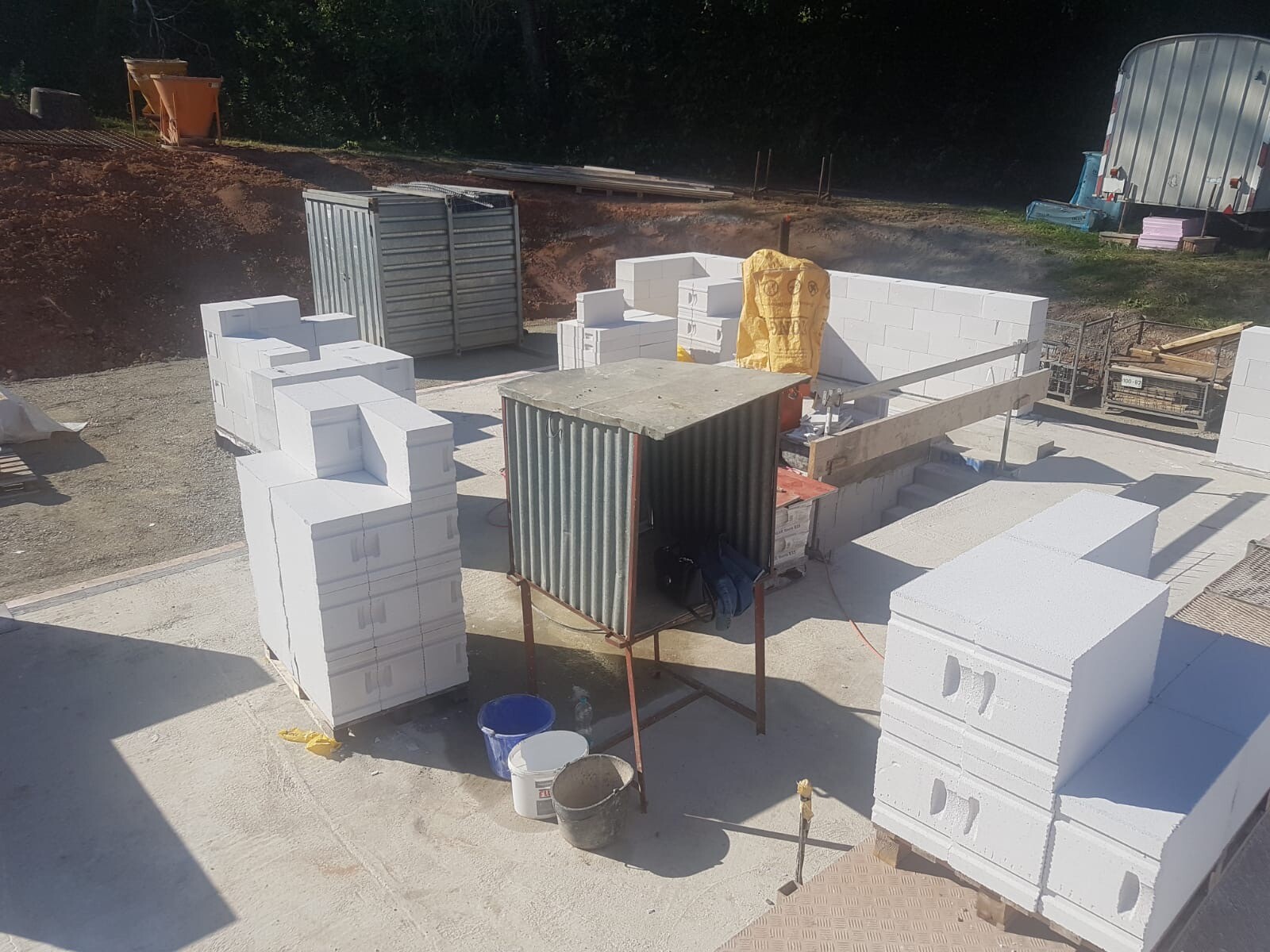 Baustelle mit weißen Styropor-Blöcken als Wände, zentrale offene Wellblech-Box, Eimer vorne