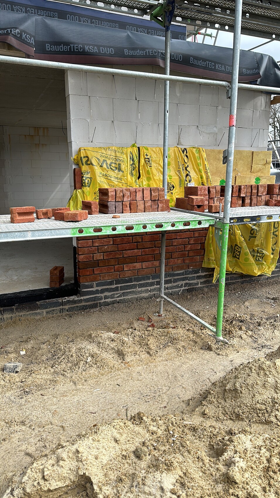 Baustelle: Ziegelsteine auf Metall-Gerüst, gelbe Dämmstoffe, unfertige Fassadenwand.