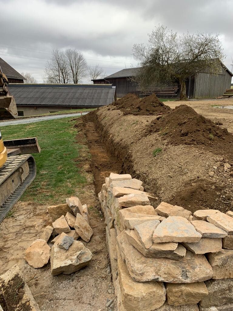 Baustelle: Erdgraben neben Steinmauer, Steine stapeln, Baufahrzeug links, Scheunen im Hintergrund.