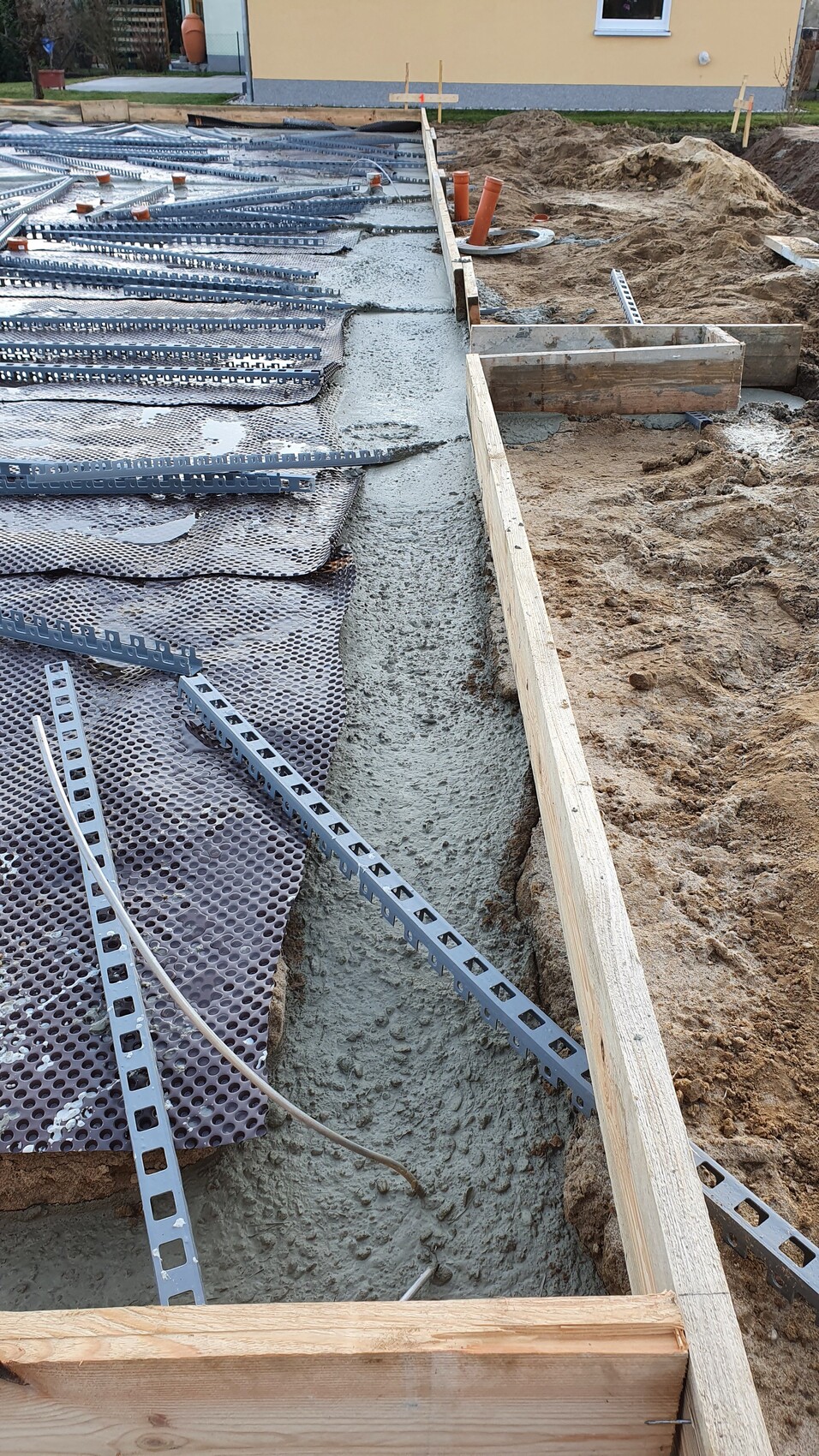Baustelle: Beton wird in Graben gegossen, Metallbewehrung, Holzformen, Sandhügel.