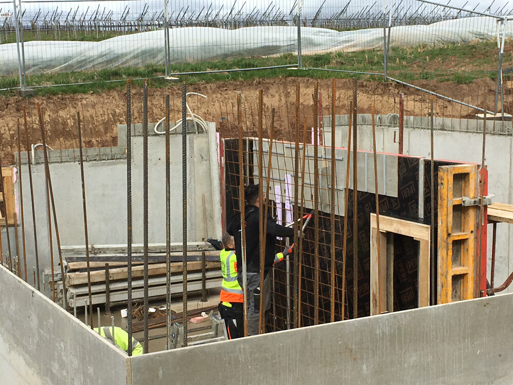 Baugrube mit Stahlbewehrung, Holzformen; zwei Arbeiter mit Warnwesten arbeiten am Fundament.
