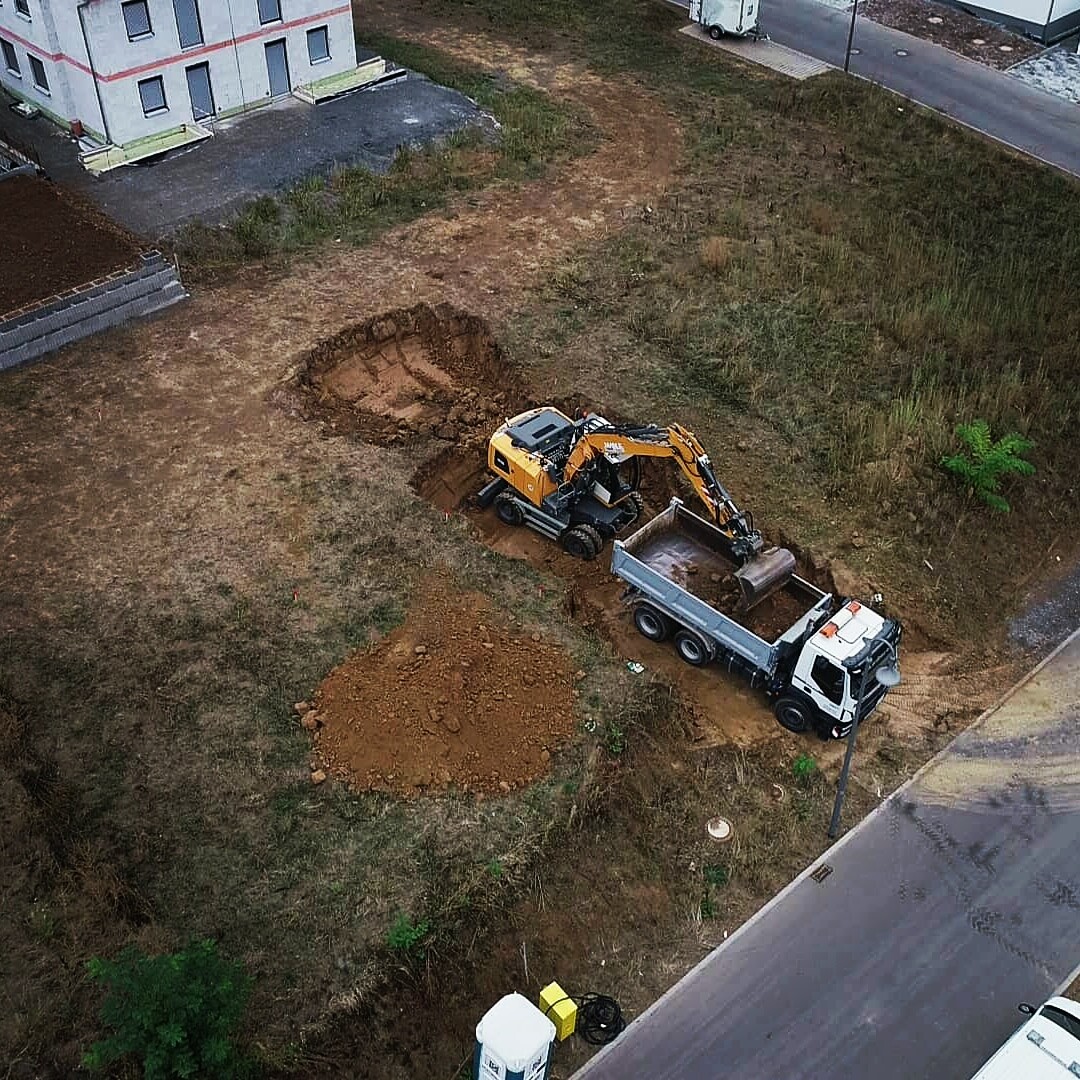 Auslegerbagger gräbt Erdreich auf Baustelle; Kipplaster lädt Bodenerde neben einem Wohnkomplex ab.