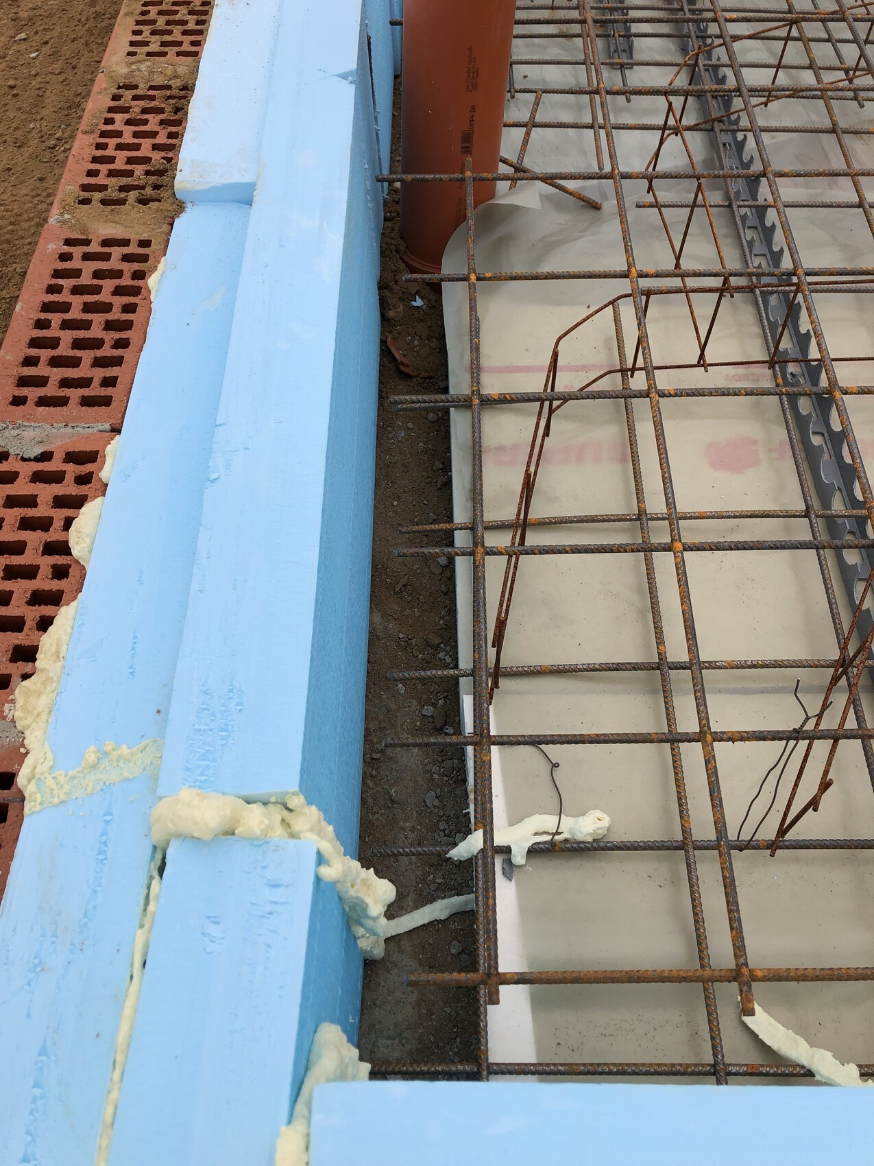 Baustelle: rostiges Bewehrungsstahlgitter, blaue Dämmung und orangefarbene Rohre neben Ziegeln.