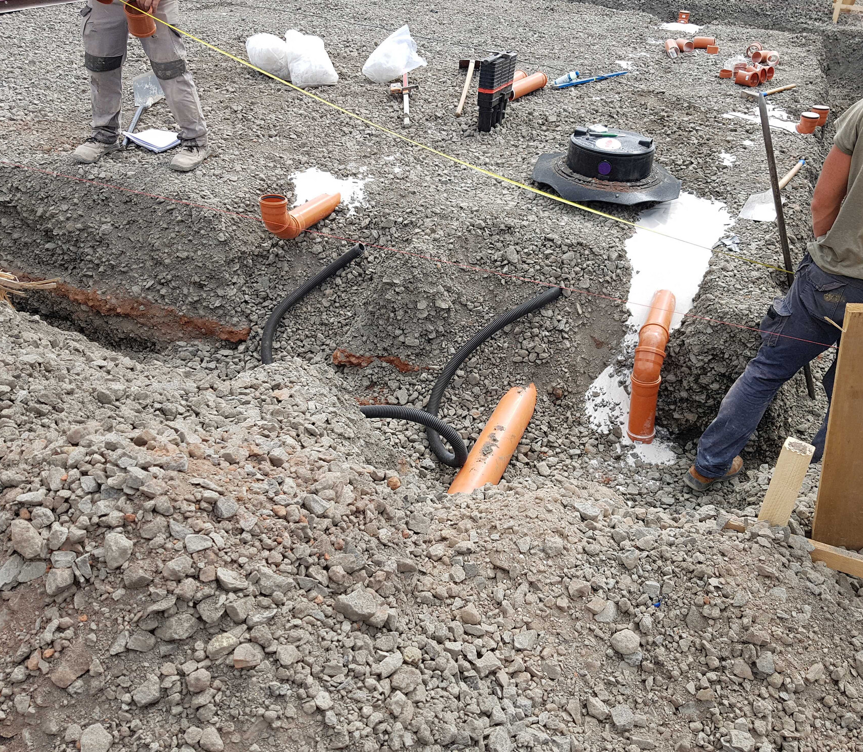 Baugrube mit orangefarbenen PVC-Rohren, schwarzen Schläuchen und Werkzeugen auf einer Baustelle.