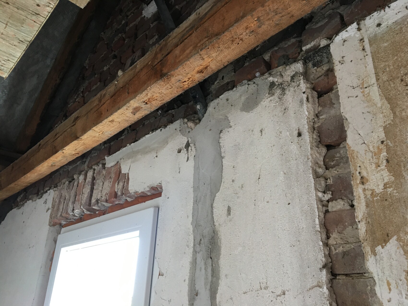 Rohbau: Ziegelwand mit Putz, Holzbalken, grobe Betonflächen und ein Fensterrahmen unten links