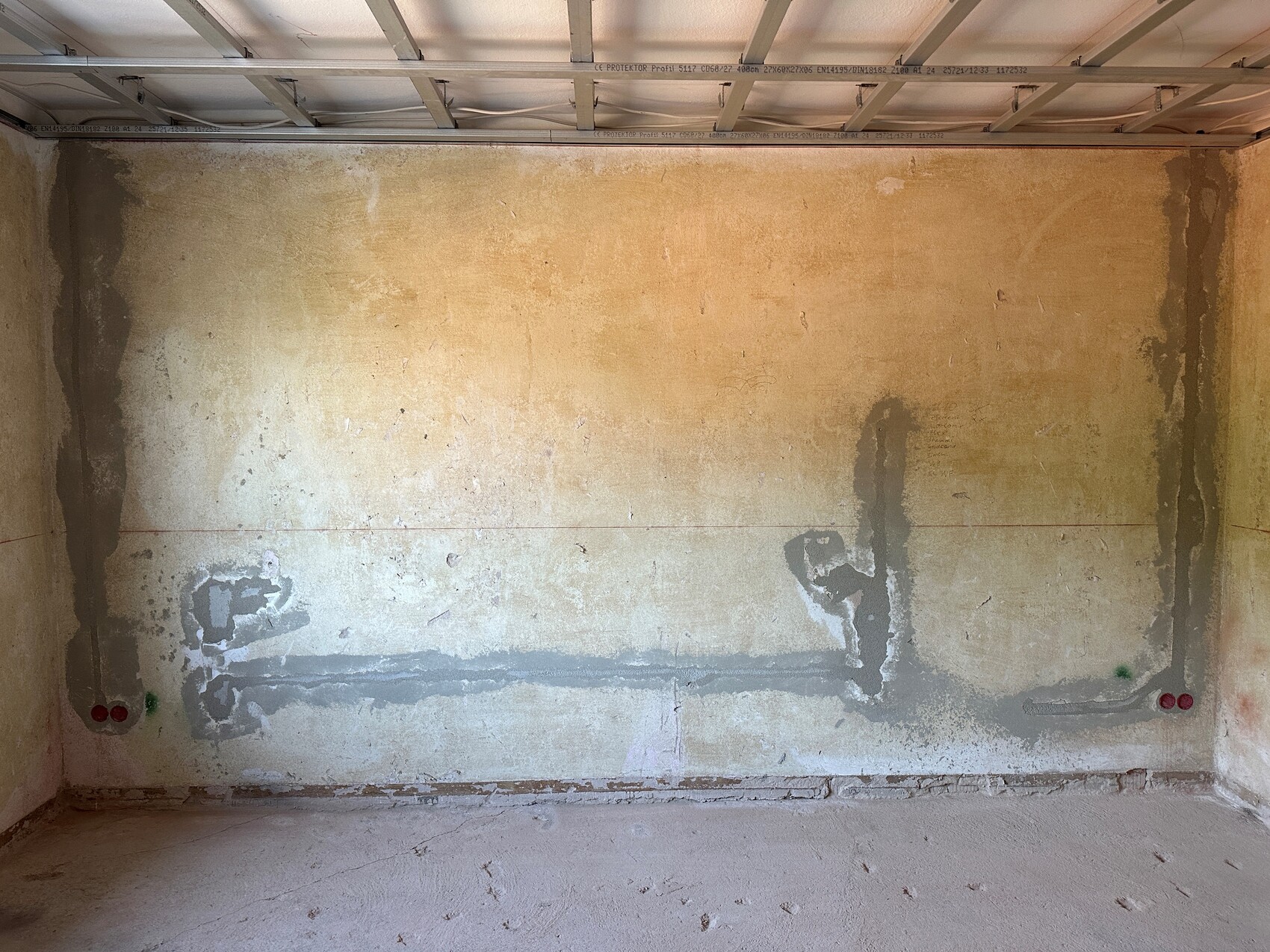 Unbearbeiteter Bau-Raum mit roher Wand, sichtbarem Decken-Trägersystem und Feuchtigkeitsspuren.