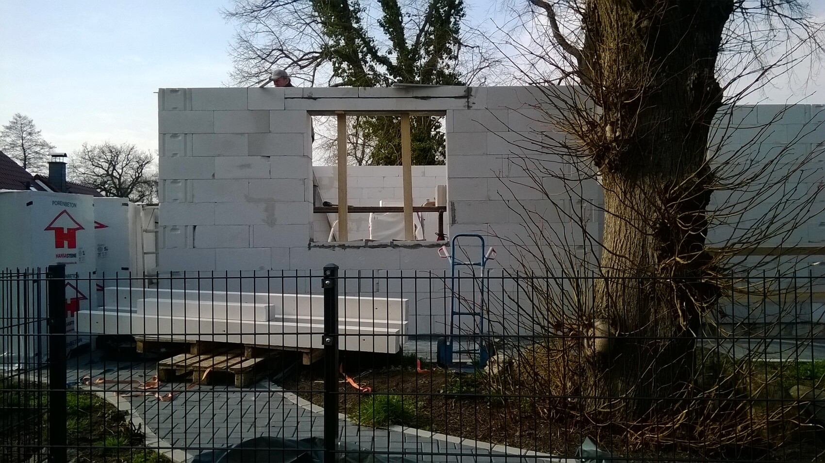 Bau einer Wand aus weißen Blöcken mit Fensteröffnung, Holzstützen, Zaun und Baum rechts.