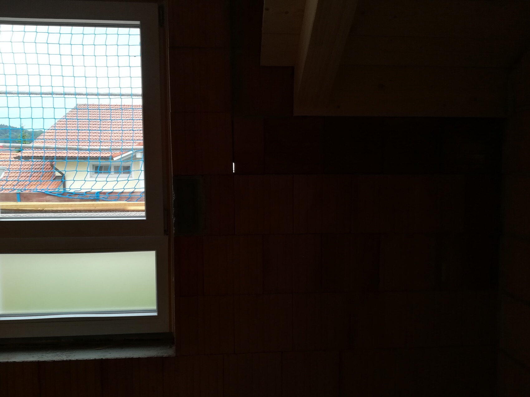 Dunkler Innenraum mit Holzwänden; Fenster mit blauem Netz, Blick auf rotes Ziegeldach.