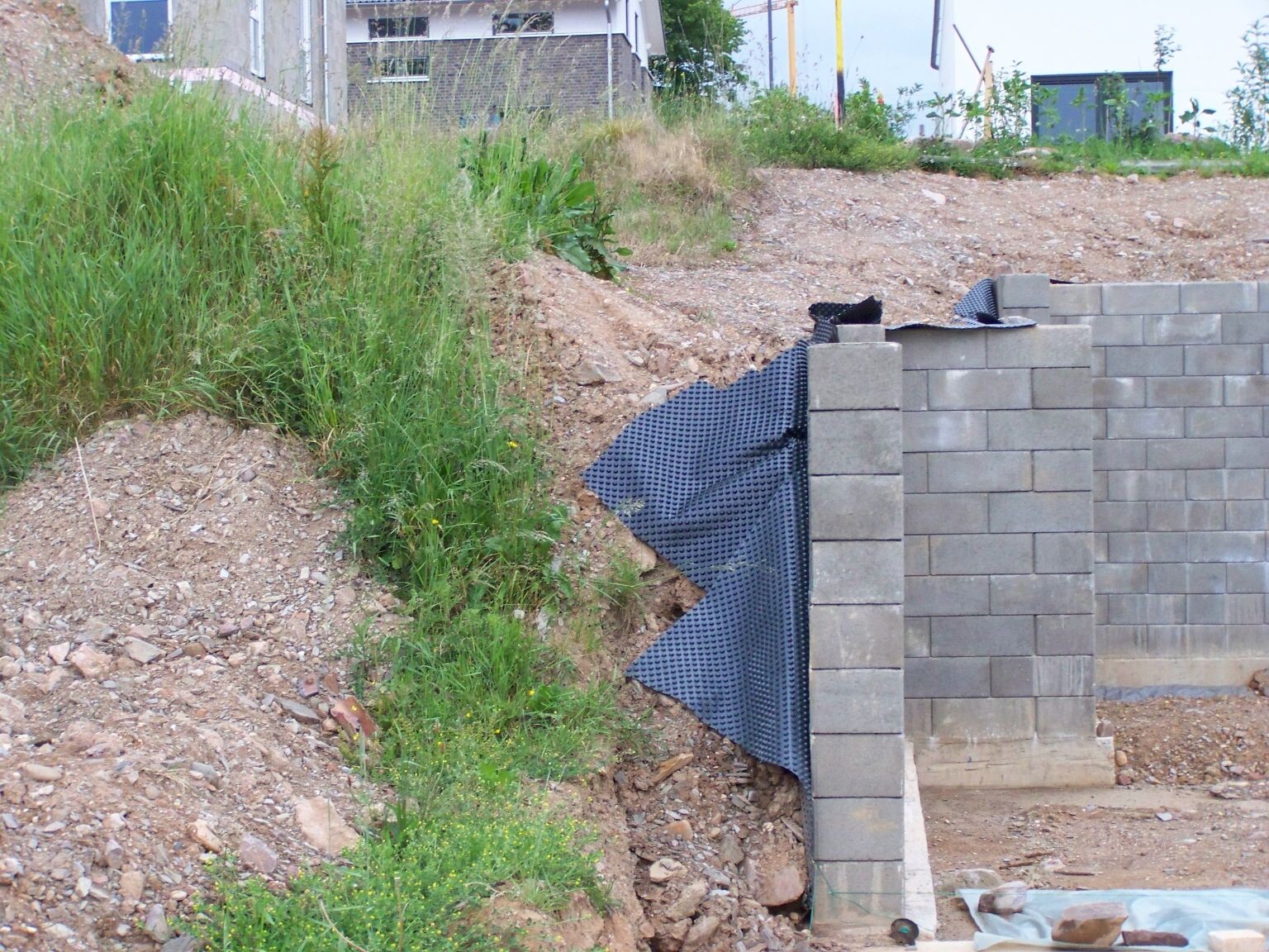 Stützwand aus grauen Betonblöcken an einer grasbewachsenen Böschung, Baugerät im Hintergrund.