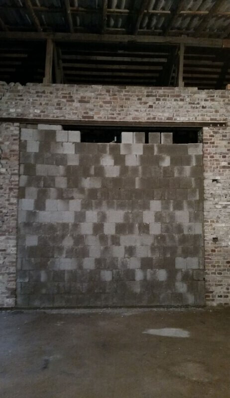 Große graue Blockwand vor einer Backsteinhalle; Decke mit sichtbaren Holzbalken.