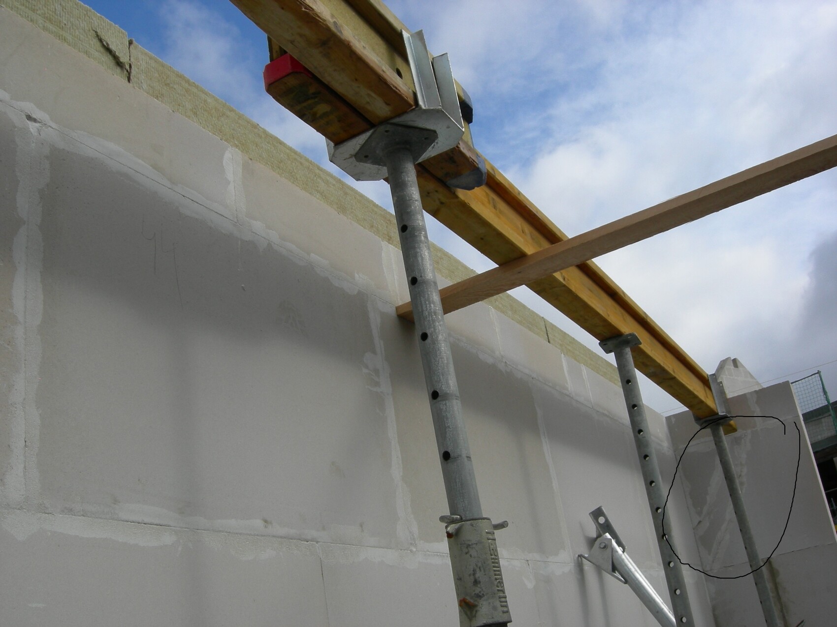 Baustelle mit Betonwand, Holzträgern und Metallgerüst unter blauem Himmel.