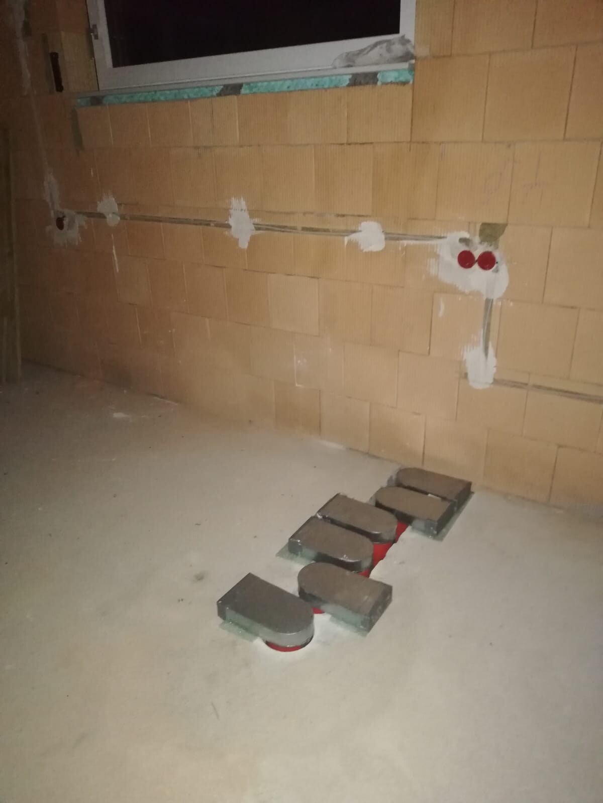 Unbearbeiteter Rohbauraum mit Ziegelwand und rohrförmigen Installationen am Boden.