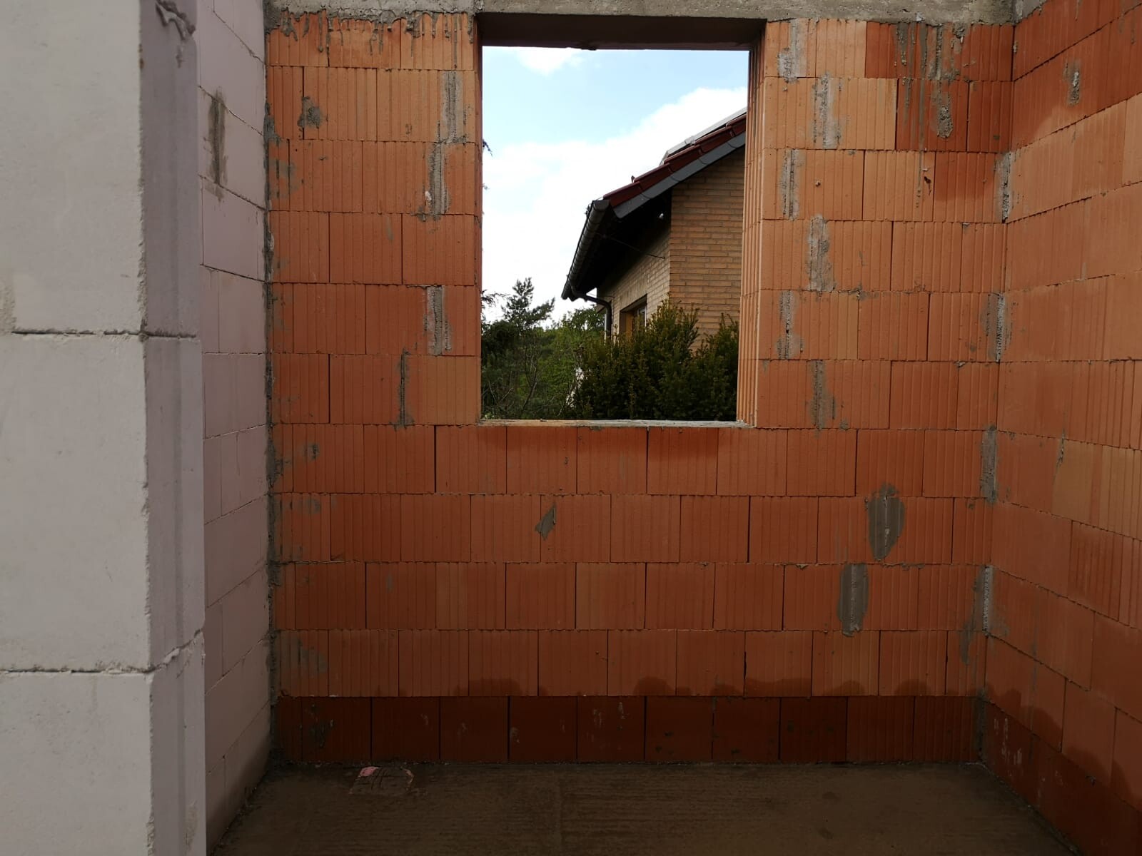 Unfertiger Raum aus roten Ziegeln mit Fensteröffnung; Blick auf Haus und Bäume dahinter.
