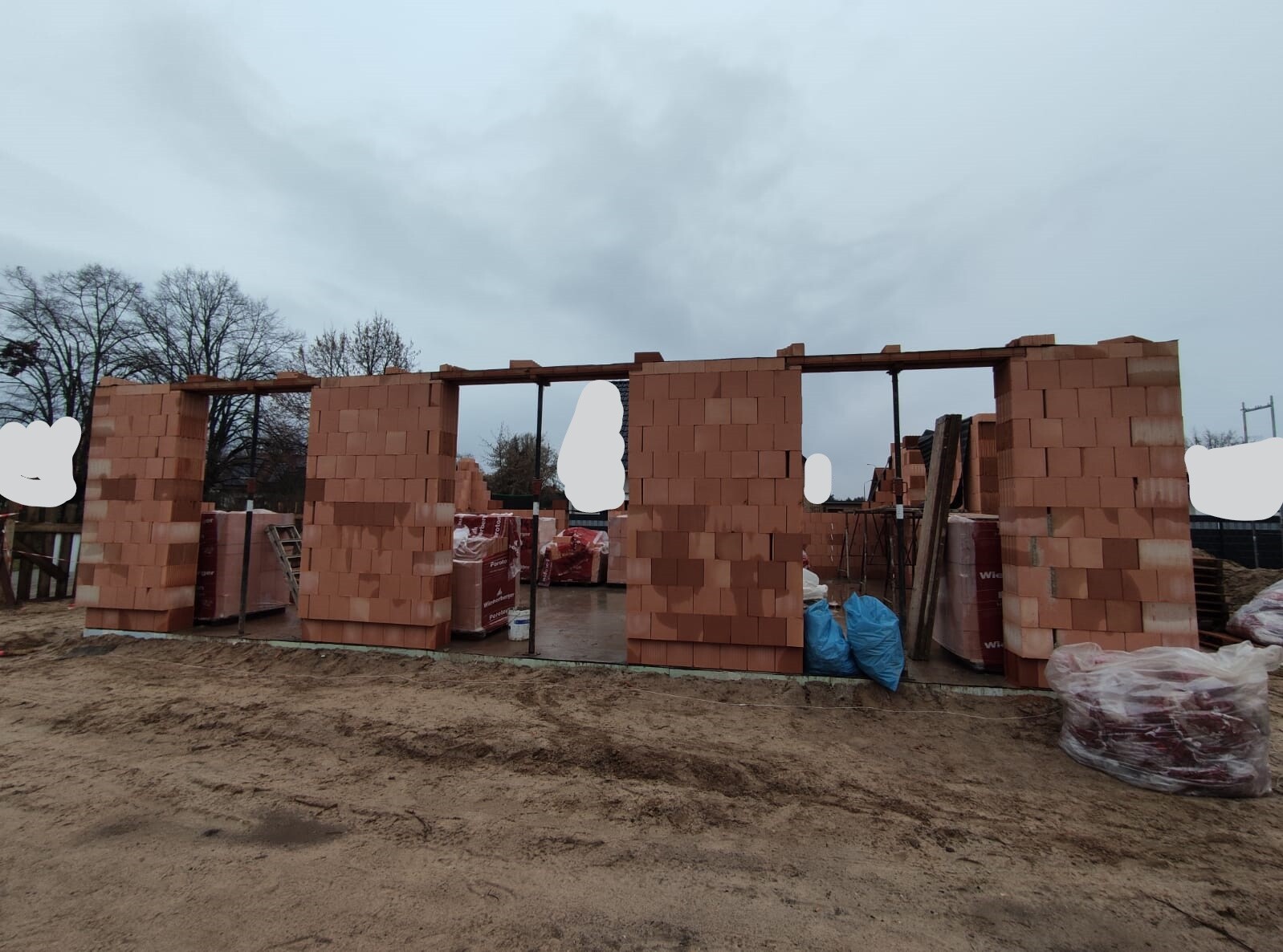 Baustelle mit unfertigen Ziegelwänden, Fensteröffnungen, Bauholz und Materialsäcke.