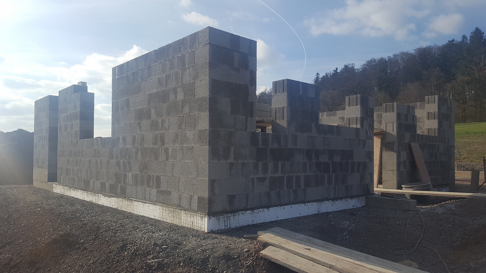 Rohbau aus grauen Betonblockwänden mit Öffnungen, Bauholz und Landschaft im Hintergrund.