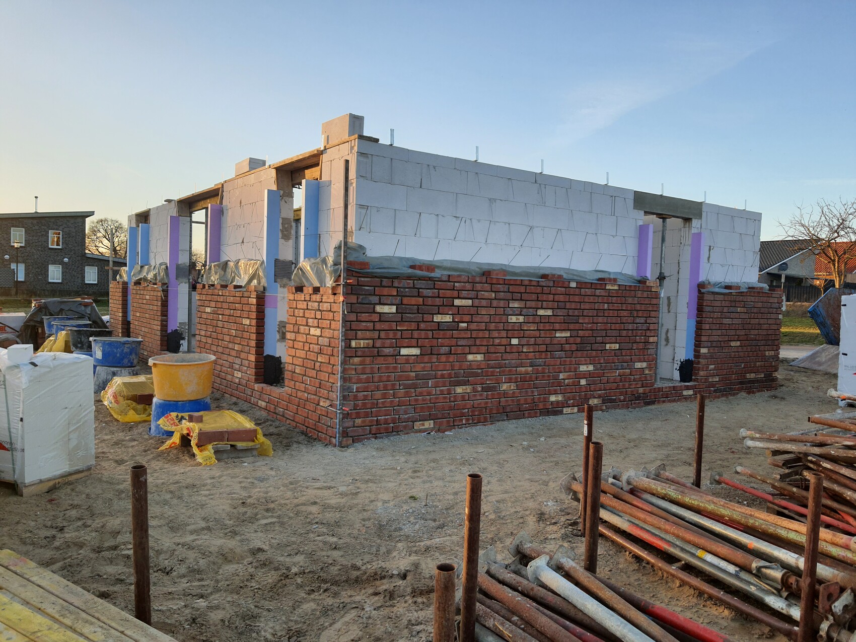 Neubau mit roten Ziegelwänden und weißem Blockaufsatz, Baugeräte und Rohrstapel im Vordergrund.