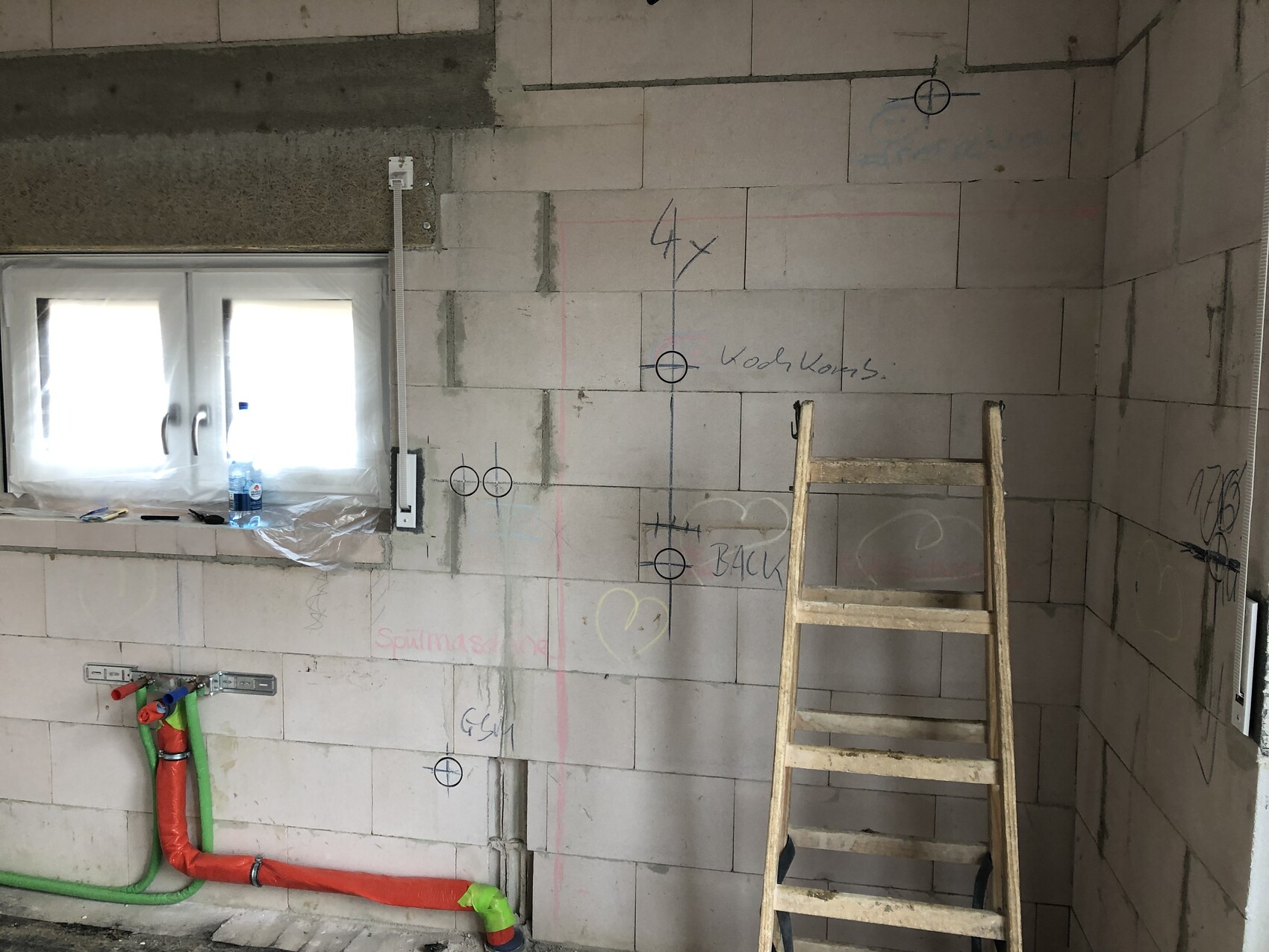 Unbearbeitete Bauwand aus Blöcken, Holzleiter, rote/grüne Rohre, Markierungen und Fenster.