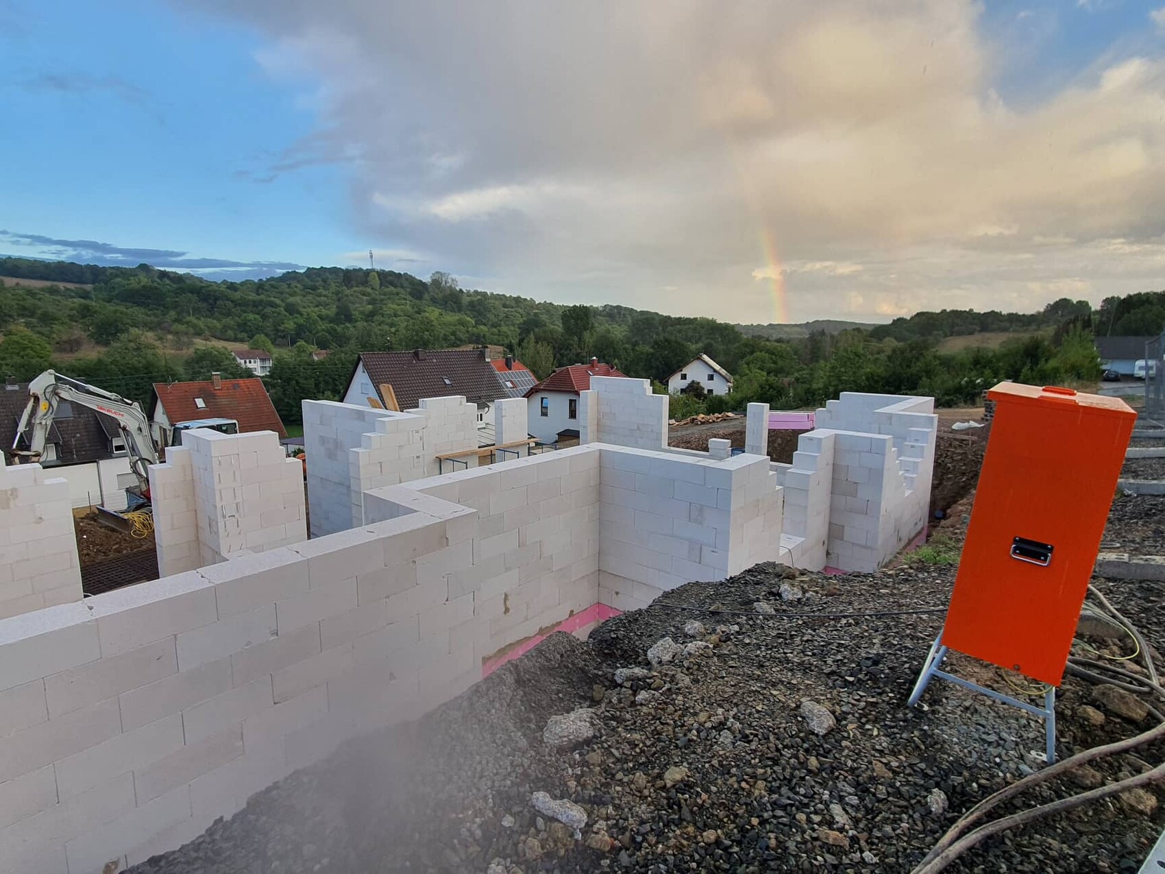 Baustelle mit weißen Betonblöcken, orange Sicherheitskasten, Regenbogen über grünem Hügelgebiet.