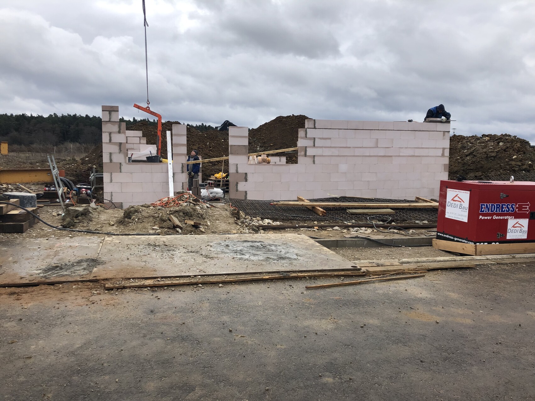 Baustelle mit halbfertiger Wand aus weißen Steinen, zwei Arbeiter, Kranhaken, rote Generatorbox.