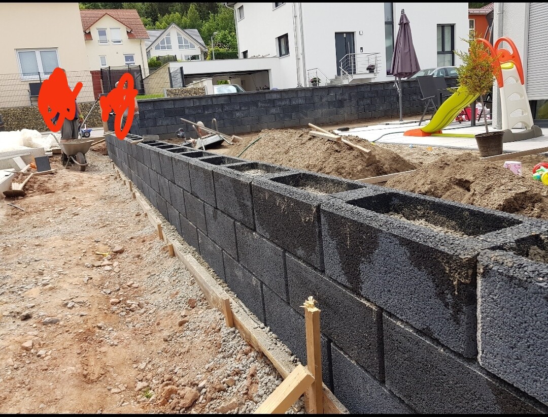 Baustelle: lange dunkelgraue Betonblock-Mauer, Erdarbeiten; Spielgerät im Hintergrund