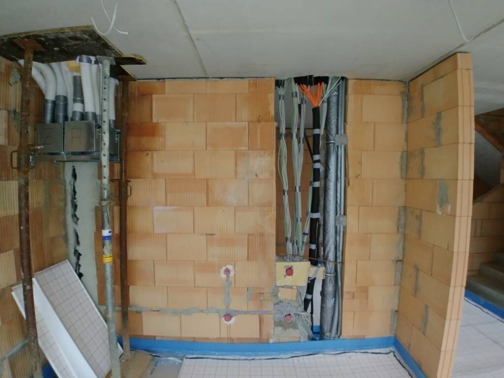 Baustellenwand aus Ziegelblöcken mit sichtbarer Elektrik- und Sanitärinstallation und Kabelbündeln.