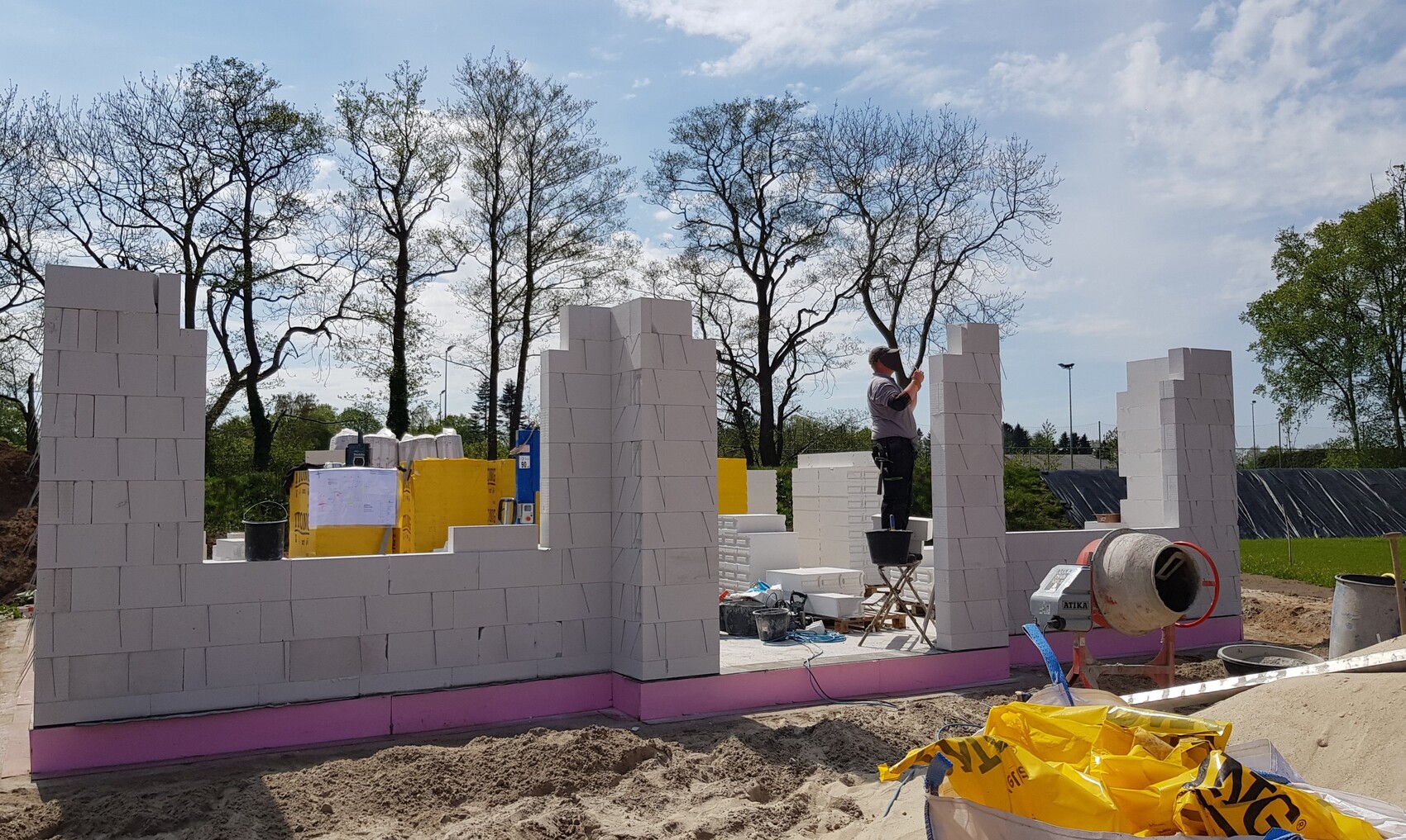 Bauarbeiter errichtet Mauer aus weißen Blöcken auf einer Baustelle; Betonmischer und Baumreihen im Hintergrund.