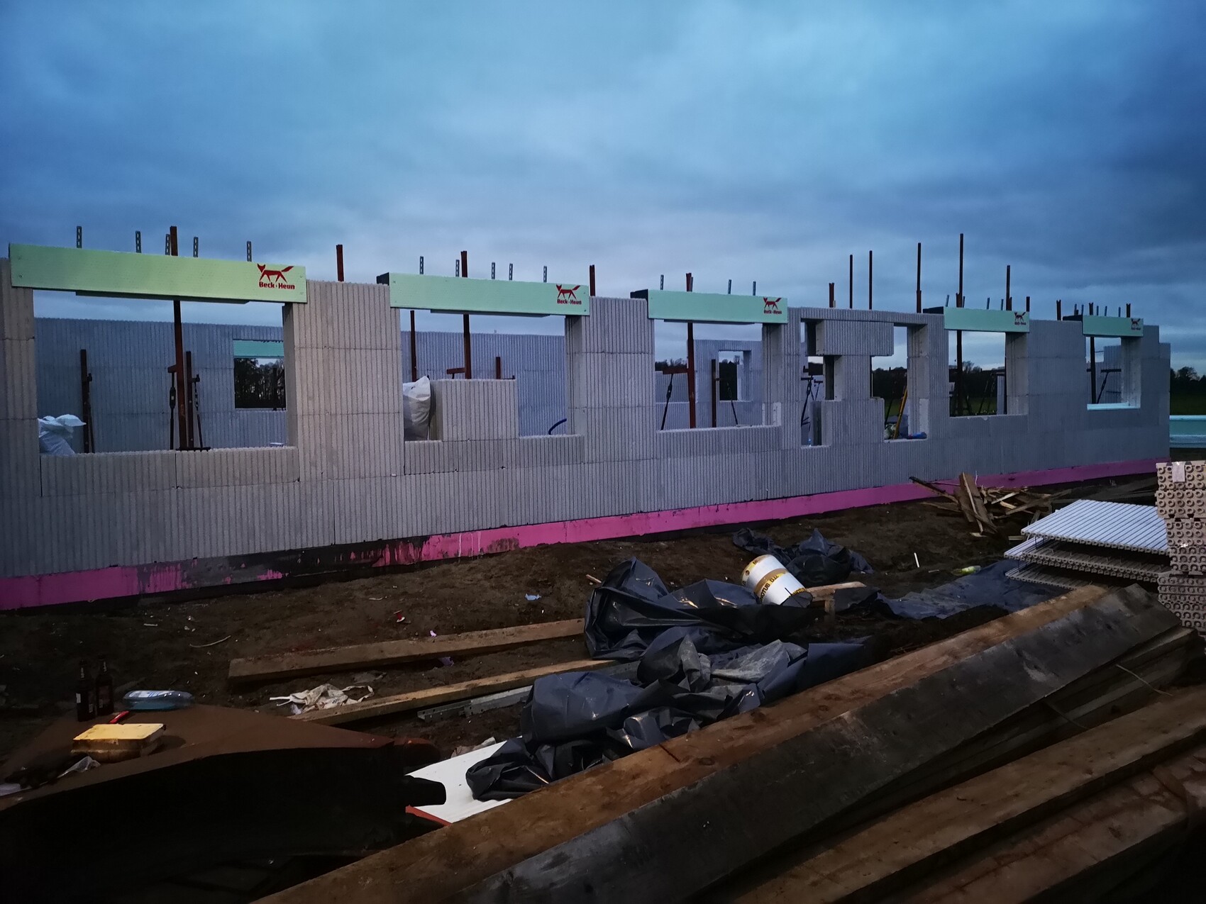 Baustelle: Betonwände mit Fensteröffnungen, Stangen ragen hoch, Holz- und Bauabfall davor.