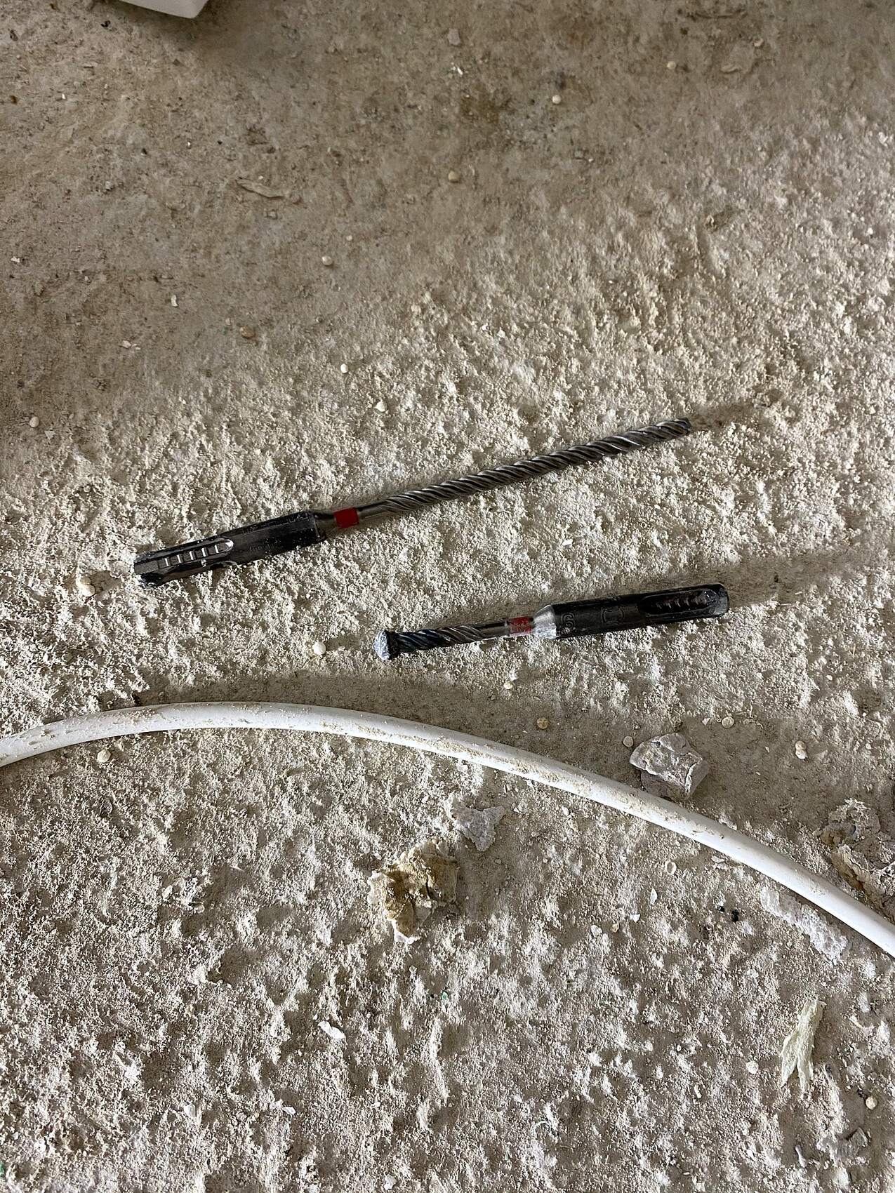 Zwei Spiralbohrer liegen auf staubigem Betonboden neben einer weißen Rohrleitung.