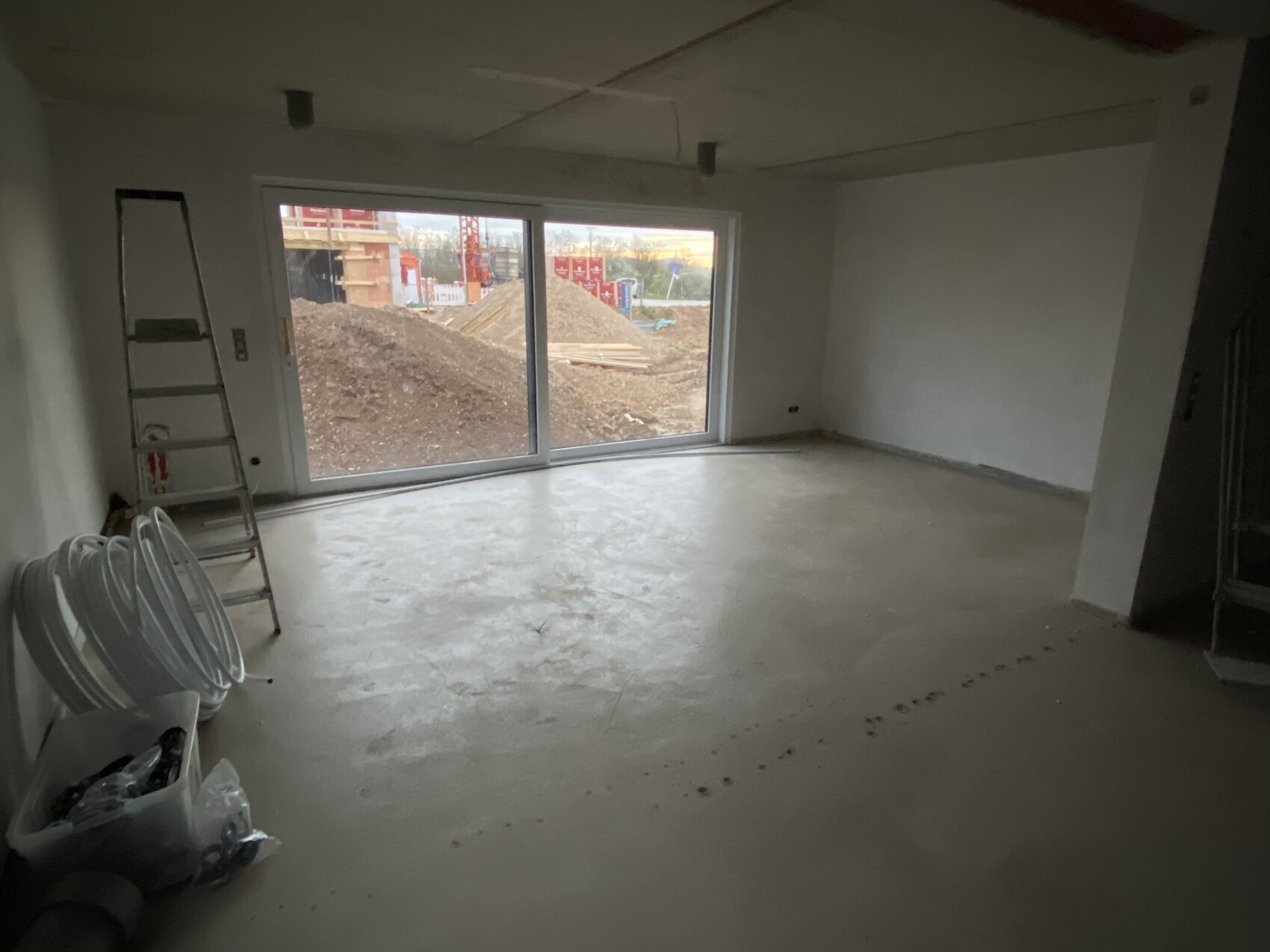 Unfertiger Innenraum: Leiter links, Schlauchrollen, Bauwerkzeuge; Baugelände hinter Glasschiebetür.