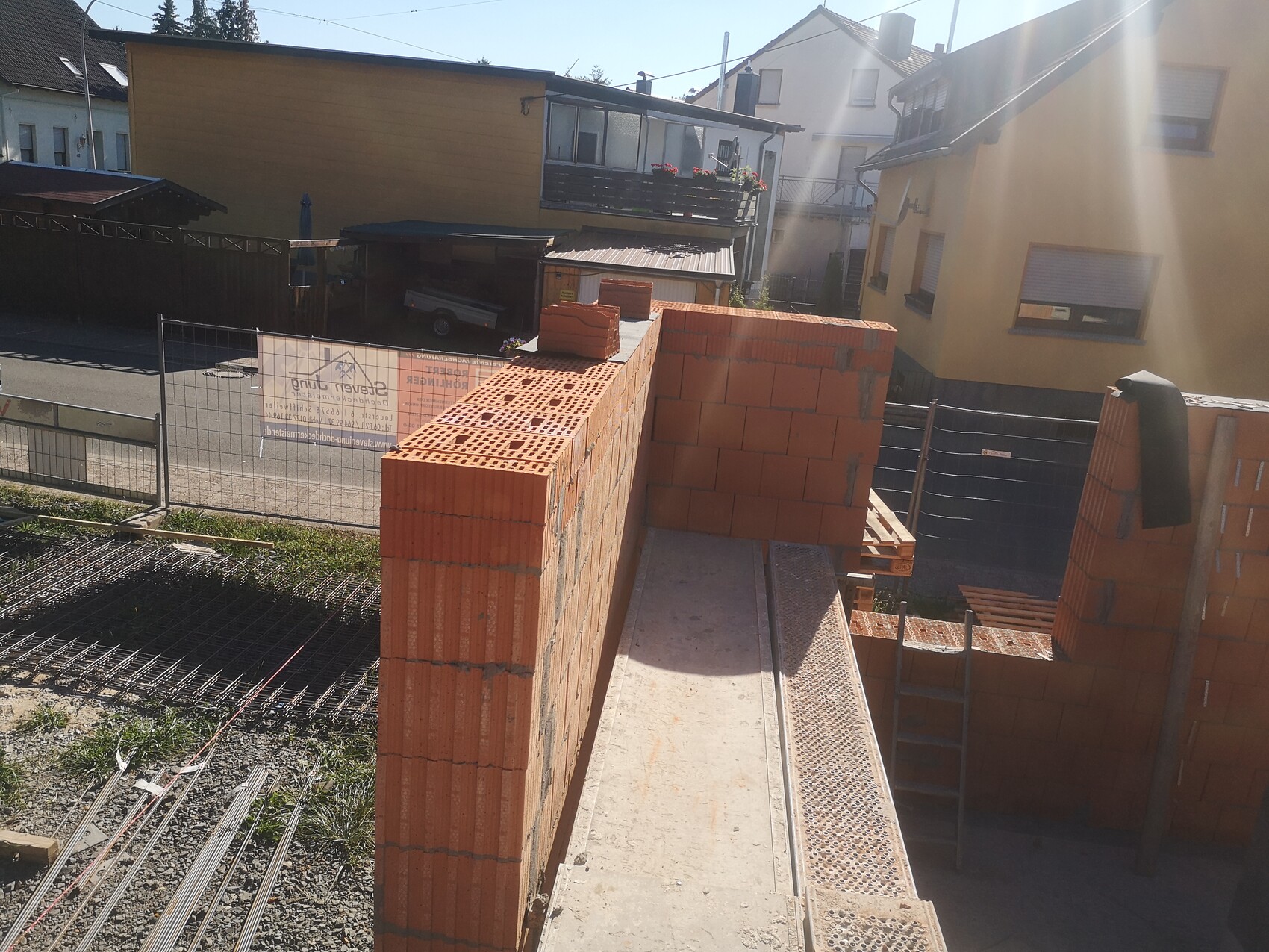 Baustelle: rote Ziegelwand im Bau, Metallplattform und Leiter, Häuser im Hintergrund.