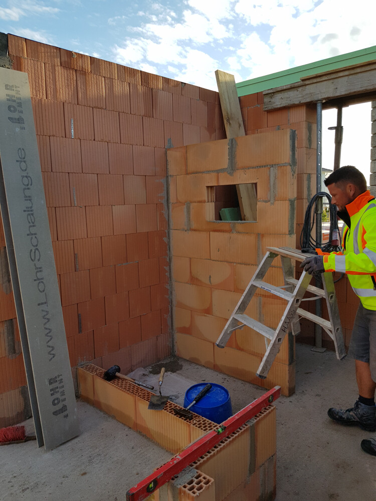 Baustelle mit orange Ziegelwand, Fensteröffnung, Arbeiter in Warnjacke neben Leiter.