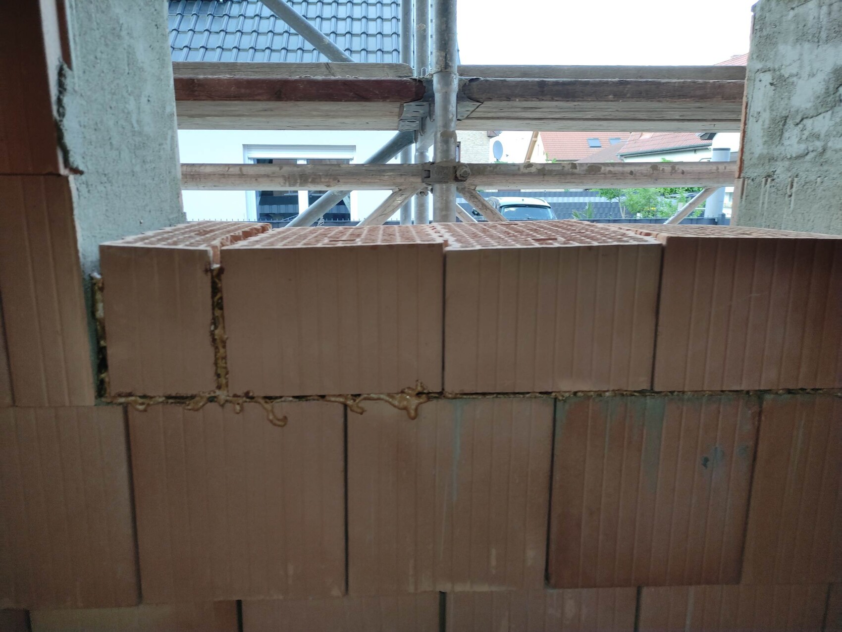 Braune Ziegelwand im Vordergrund einer Baustelle mit Gerüst, Häusern im Hintergrund.