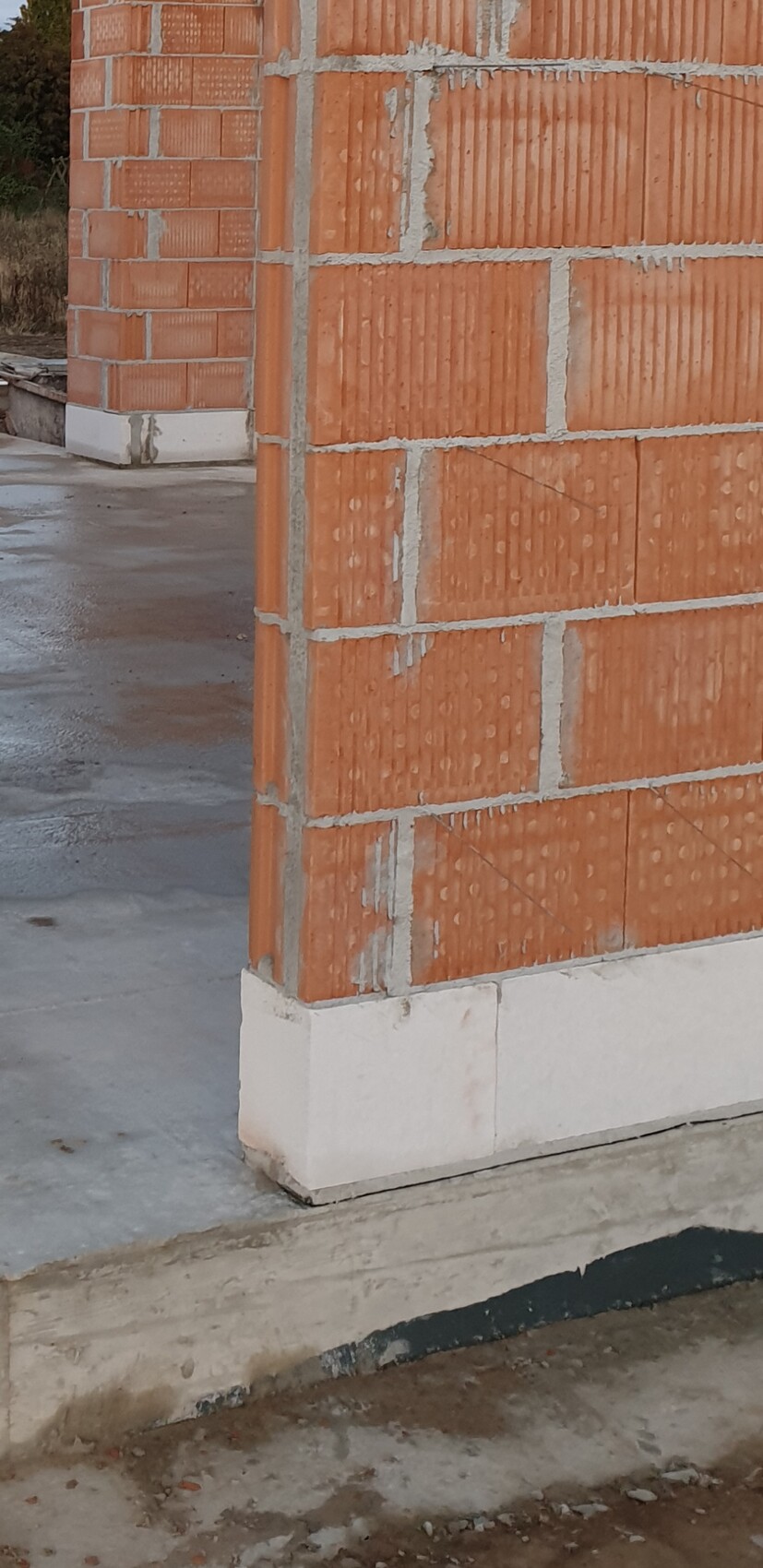 Eckansicht eines Rohbaus mit orangefarbenen Ziegelwänden, weißem Fundament und nassem Boden.